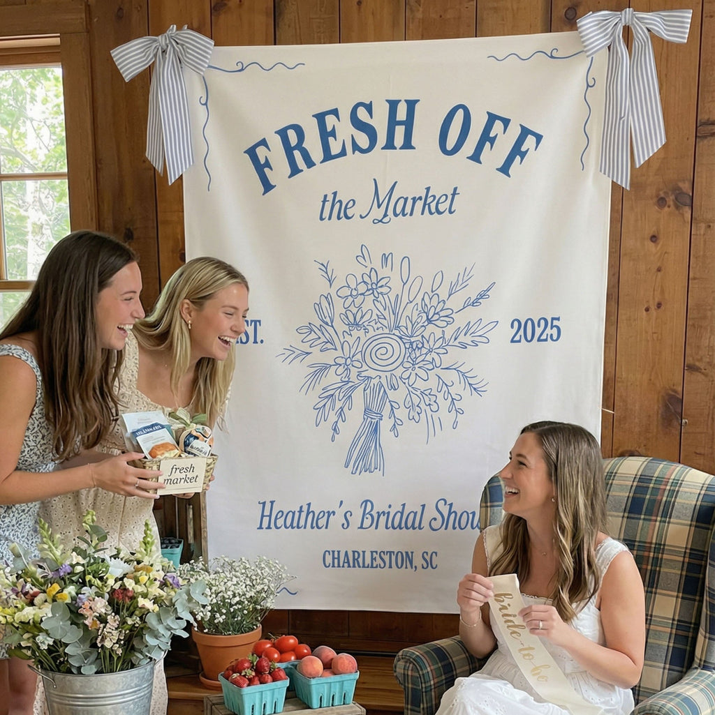 Two women standing and one sitting in a room with a 'Fresh Off the Market' banner and floral arrangements.