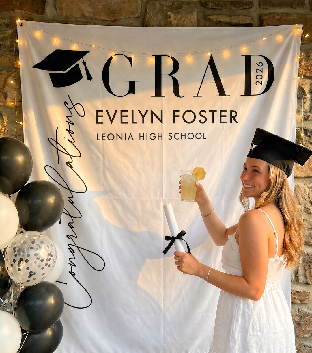 Graduating high school senior holding a drink in front of a 'GRAD' banner with personalized details.